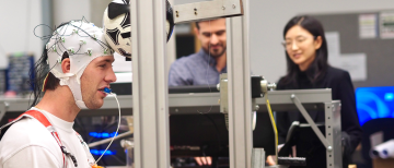 A male participant performs a controlled soccer header in a lab, wearing an EEG cap and custom mouthguard to track brain activity and head movement, while two researchers observe in the background.