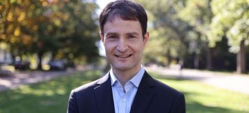 Seaspan Chair Dr. Adrien Desjardins stands on Main Mall at UBC Vancouver Campus. He smiles at the camera, wearing a dark blazer and light collared shirt.